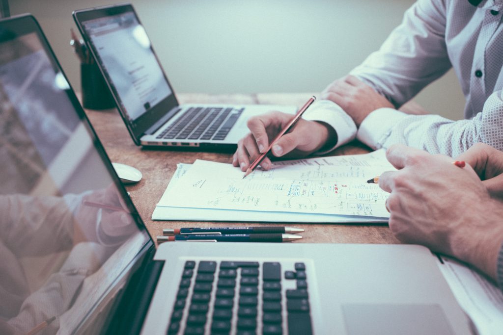 Two people sit at a table with two laptops and pens and paper.