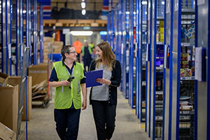 A smiling manager speaks to a happy warehouse employee while they walk through the warehouse.