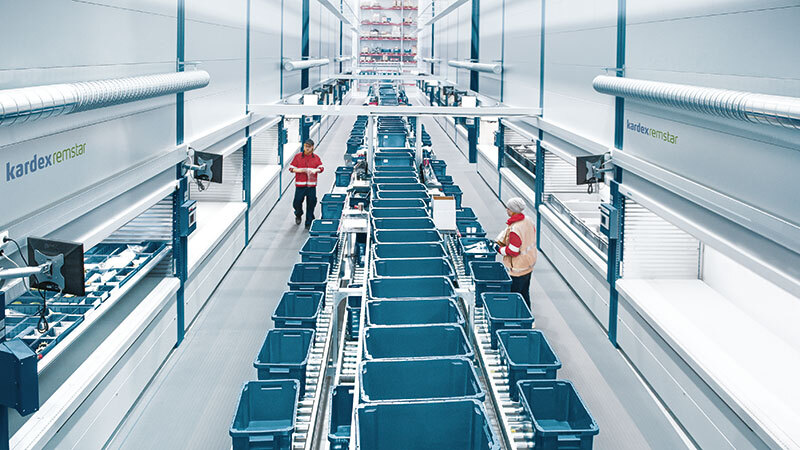 Two employees pick and pull from an aisle of Kardex VLMs with conveyor running down the middle